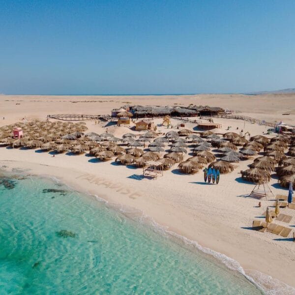 An aerial view of Paradise Island beach in Hurghada featuring crystal clear turquoise waters, white sands, and traditional thatched umbrellas