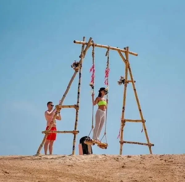 Tourists enjoying a tall wooden swing on the sandy beach of Paradise Island in Hurghada under a clear blue sky.
