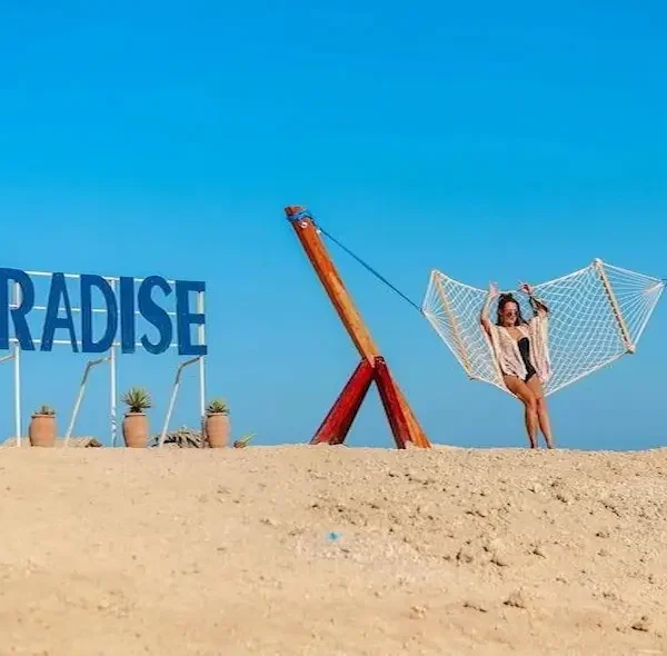 A large blue "PARADISE" sign and a wooden hammock on a sandy beach with a woman posing at Paradise Island Hurghada.