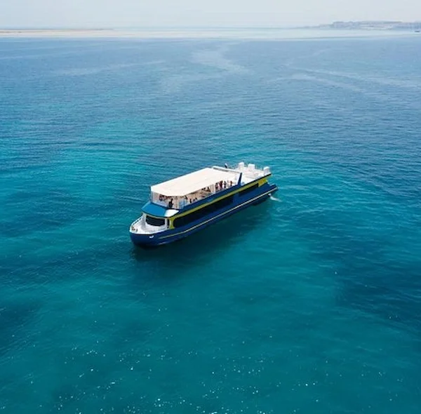 An aerial drone shot of a blue and yellow semi-submarine boat cruising through clear turquoise Red Sea water.