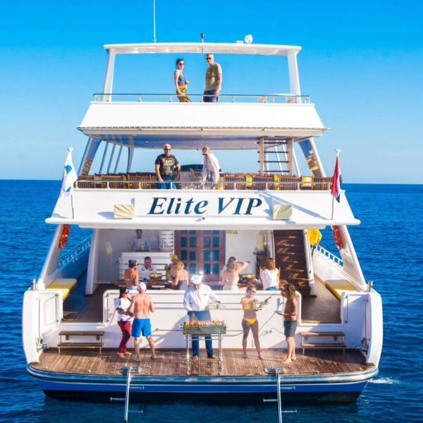 A wide rear view of the Elite VIP luxury boat in Hurghada featuring tourists enjoying a live BBQ lunch on the wooden deck amidst the crystal-clear blue waters of the Red Sea