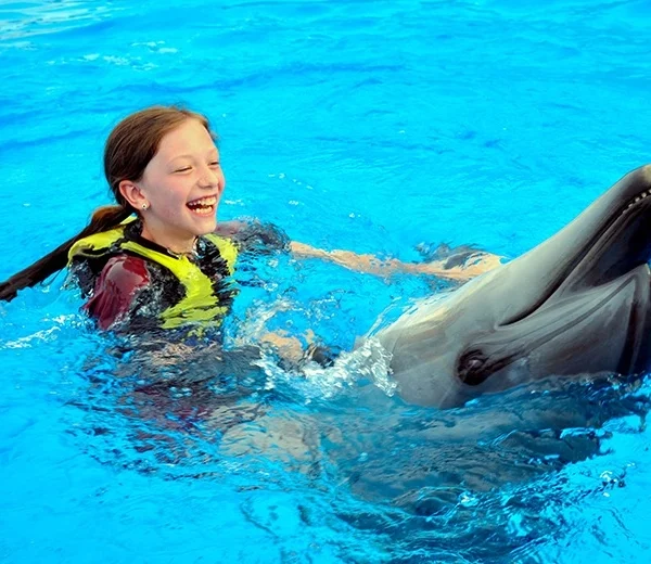 A young girl with long dark hair wearing a yellow life vest, smiling while swimming alongside a dolphin.