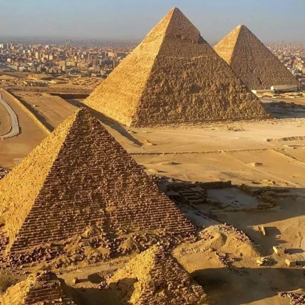 A high-angle, aerial view of the three main Pyramids of Giza in the desert, with the dense urban cityscape of Cairo stretching into the horizon behind them