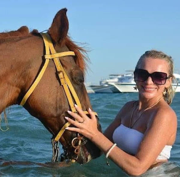 A woman wearing sunglasses smiling while standing in the Red Sea with a brown horse wearing a yellow harness.
