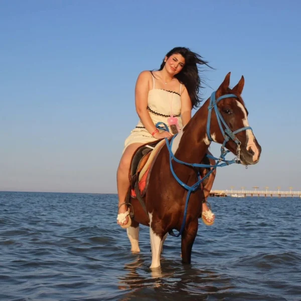 smiling woman wearing sunglasses and a white top standing in the sea while gently petting a brown horse.