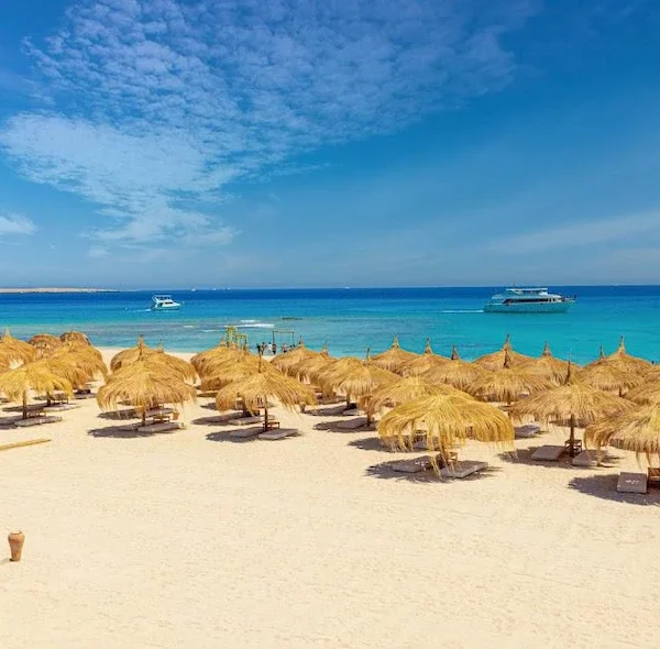 A panoramic view of Hula Hula Island beach featuring numerous straw umbrellas on white sand, turquoise water, and several boats in the distance.