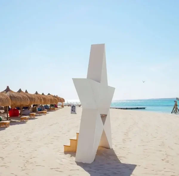 A large white star-shaped decorative structure on the white sandy beach of Hula Hula Island with straw umbrellas and turquoise water.