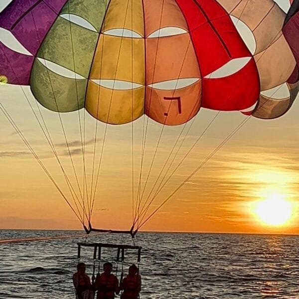 Three people parasailing together at sunset in Hurghada, with the golden sun reflecting on the Red Sea waves under a large rainbow parachute