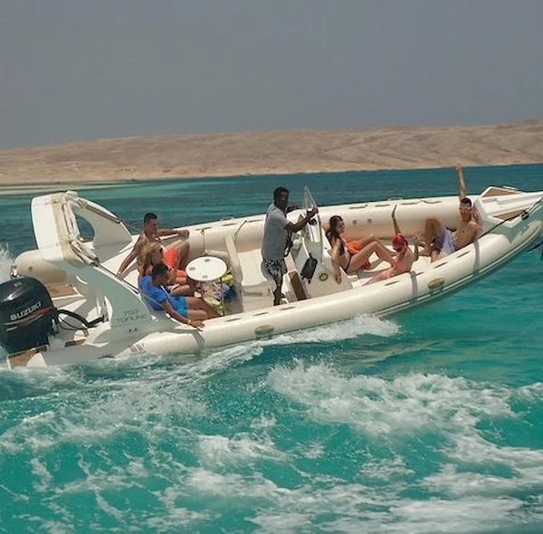 A group of people cheering on a cream-colored RIB speedboat as it cruises through turquoise water near a desert coastline.