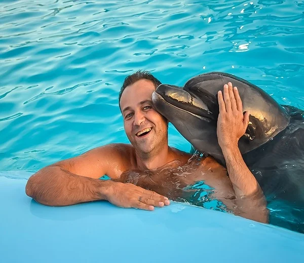 A man smiling and leaning against the edge of a pool while a dolphin gently touches its snout to his cheek