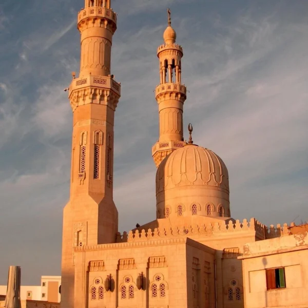 Two tall, ornate stone minarets and a central dome of a mosque glowing in the warm light of a sunset against a blue sky