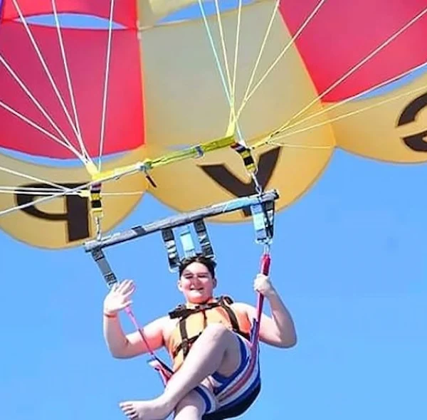 A person in a life jacket waving happily while parasailing under a multi-colored red, yellow, and purple parachute against a clear sky.
