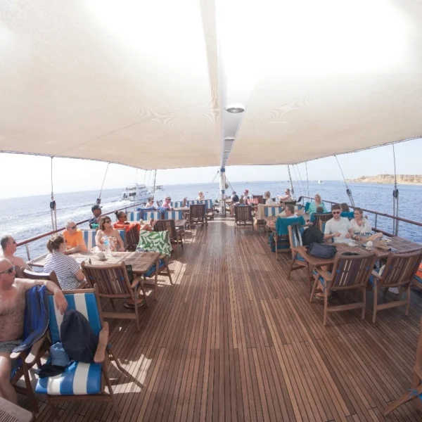 Tourists seated at wooden tables under a large canopy on the spacious deck of a luxury sailing ship.