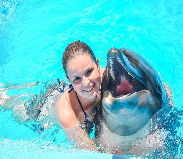 A smiling woman in the water holding a dolphin that has its mouth open in a playful expression.