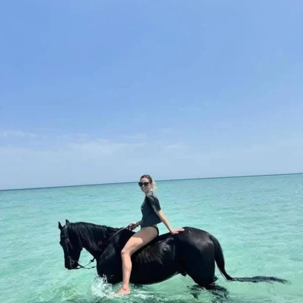 A woman in a black swimsuit riding a black horse through bright turquoise ocean water under a clear sky.