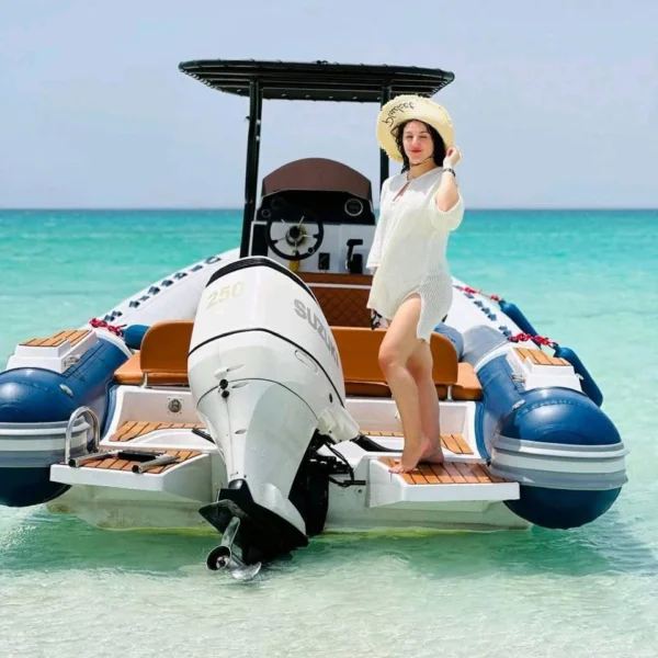 A woman in a white cover-up and sun hat stands on the rear deck of a blue and white RIB boat in shallow turquoise water.