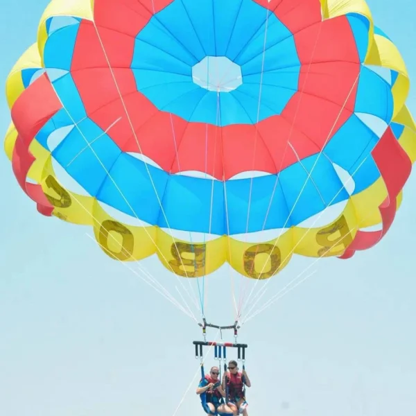 Two people in red life vests tandem parasailing under a large blue, red, and yellow bullseye-patterned parachute against a pale blue sky.