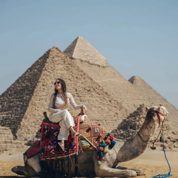 A woman in white clothing sitting on a decorated camel in the desert, with the three Great Pyramids of Giza blurred in the background.
