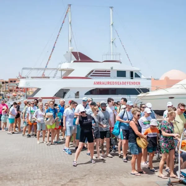 A long queue of tourists boarding a large white and red excursion boat named Amral Red Sea Defender at a sunny dock.