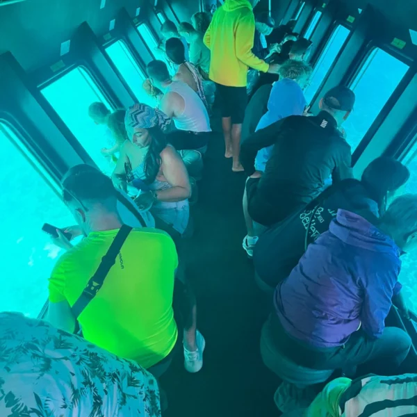An interior view of a semi-submarine filled with tourists looking through large glass windows at the turquoise underwater ocean.