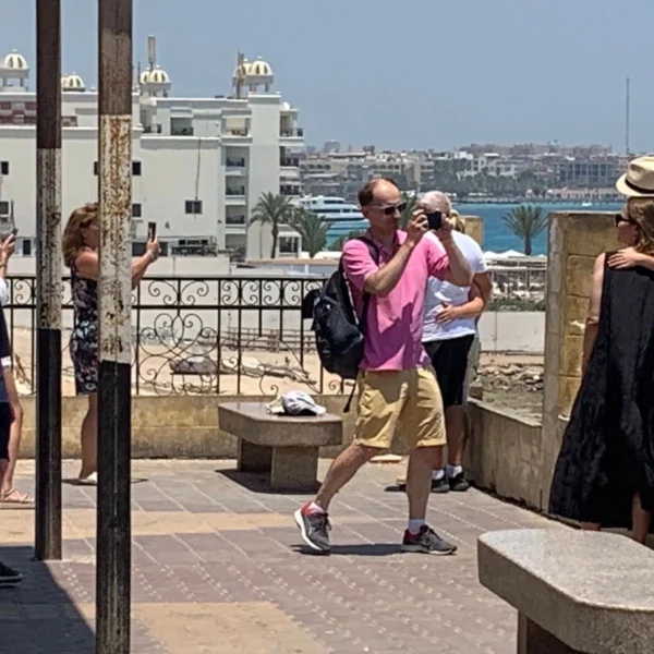A group of tourists on a sunny terrace overlooking a coastal city with white buildings and blue water, taking many photos
